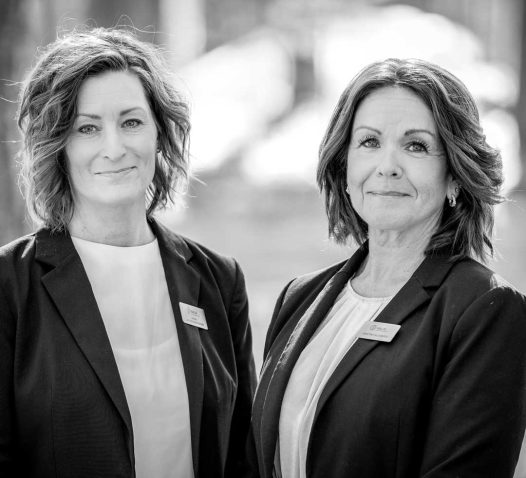 Two women in business attire stand side by side, smiling slightly, with name badges on their jackets, in a black and white photograph.