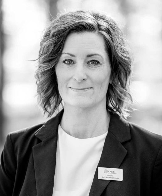 A woman with shoulder-length hair smiles slightly, wearing a blazer and a name tag that reads "KALIX Ulrika Jacobsson Olsson," set against a blurred outdoor background.